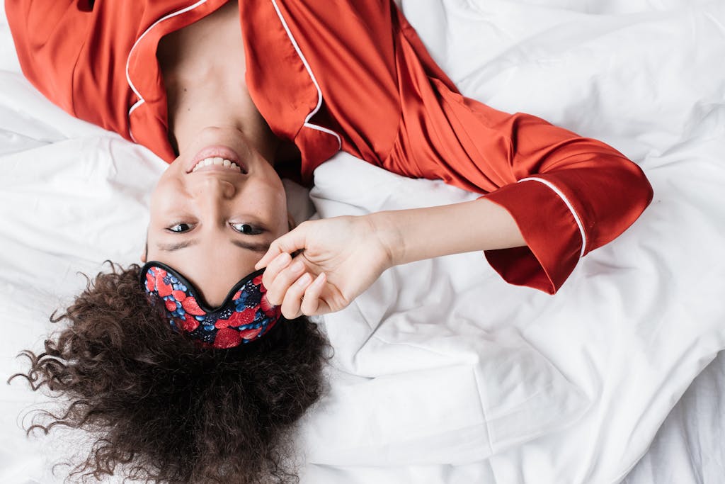 Happy woman in red sleepwear smiling while adjusting her sleep mask. Top view.