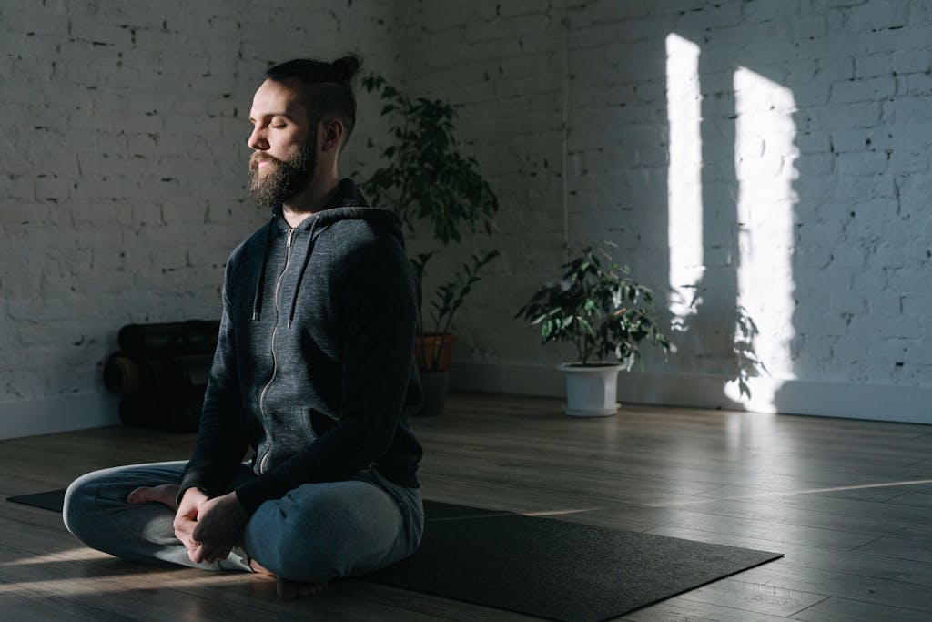 A man in a hoodie meditates in the lotus position on a yoga mat inside a bright room using best meditation devices
